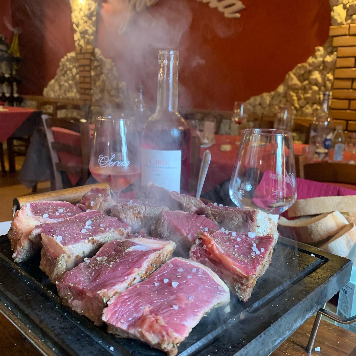 Bistecche crude, spesse e fumanti, condite con sale e cotte su una griglia di pietra rovente, servite in un ristorante con del vino.