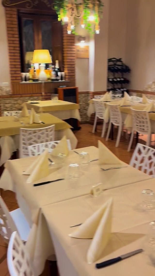 La sala da pranzo di un ristorante è arredata con diversi tavoli coperti da tovaglie beige, sedie bianche e tovaglioli piegati.