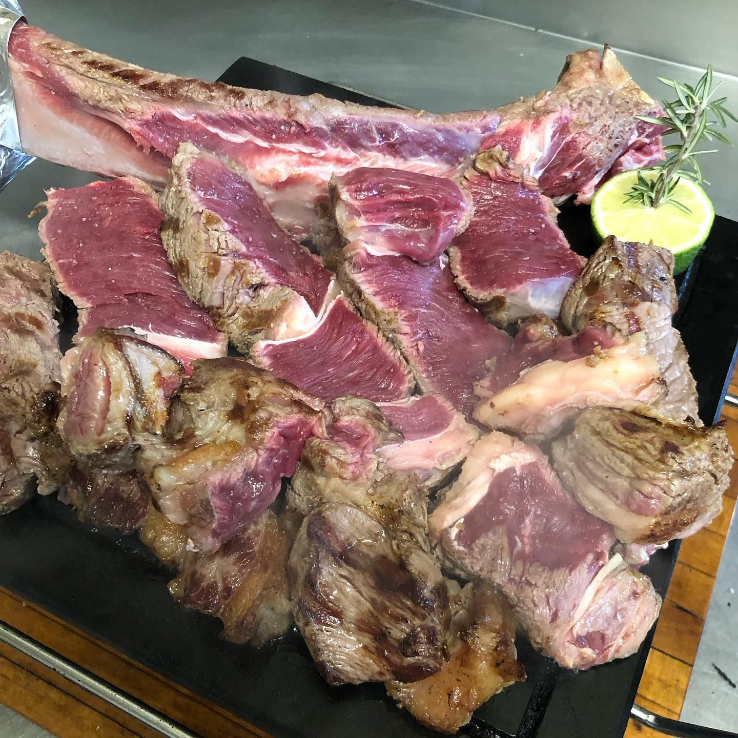 Una grande bistecca tomahawk tagliata a fette, servita su un piatto di pietra scura e guarnita con mezzo lime e un rametto di rosmarino.