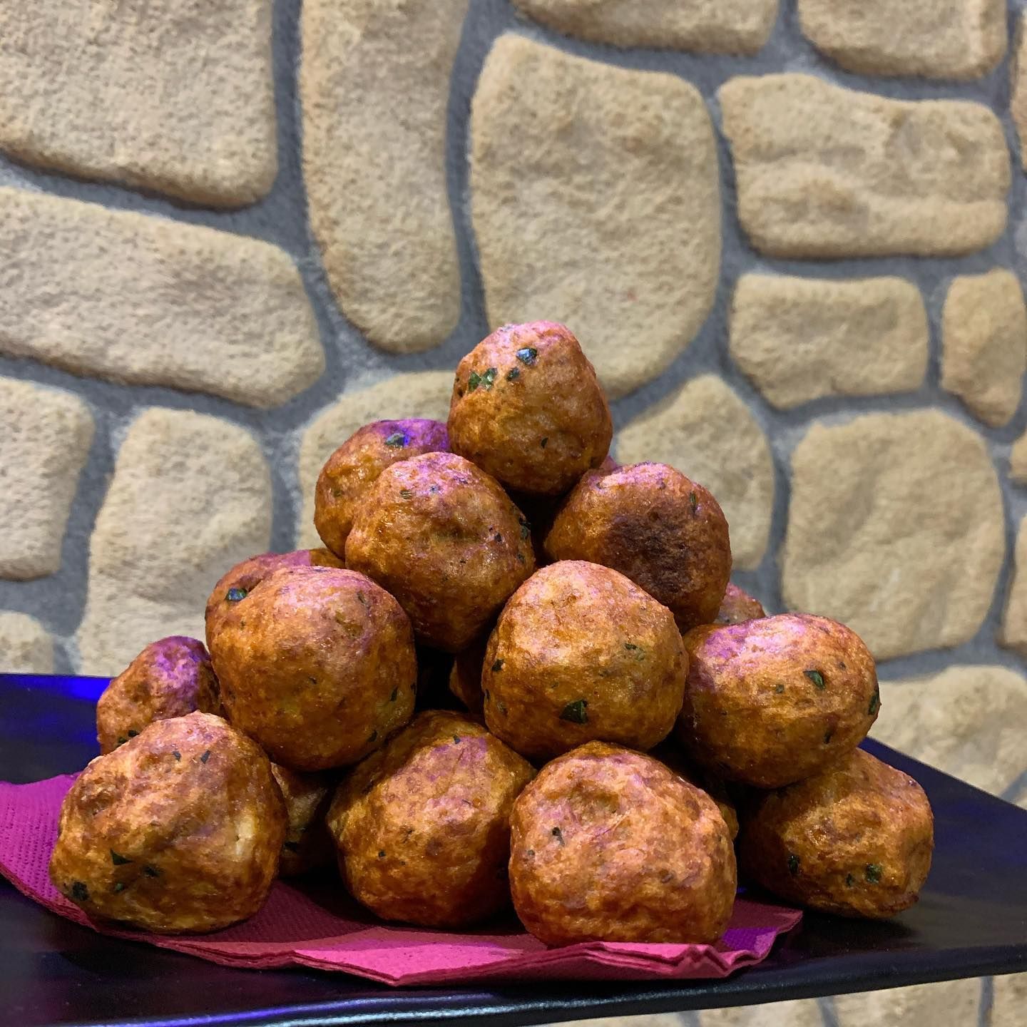 Una piramide di polpette fritte dorate, servite su un tovagliolo rosa, sullo sfondo di un muro di pietra dalla texture particolare.