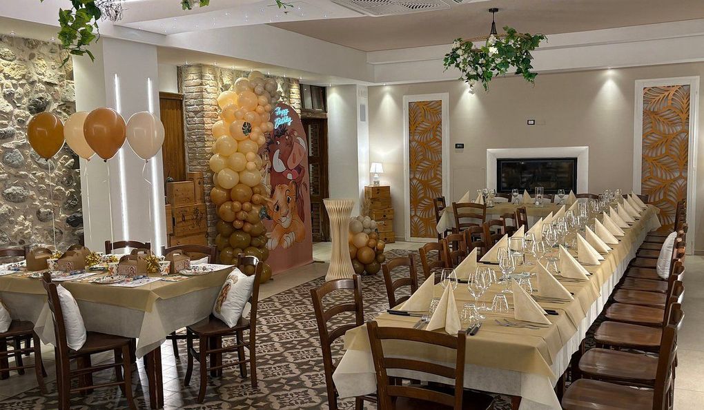 Uno spazio per eventi decorato con lunghi tavoli apparecchiati per la cena, palloncini color beige e uno sfondo di muro in pietra dai toni neutri.