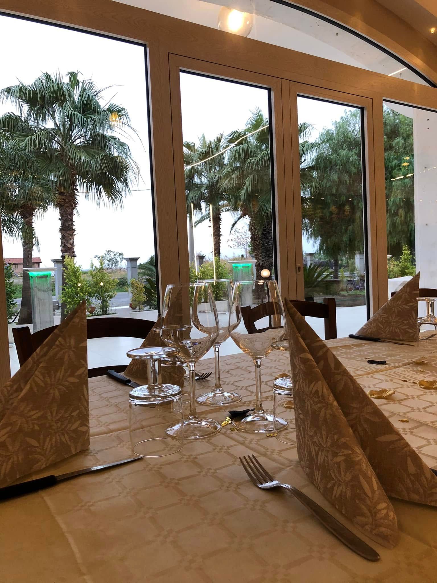 Un tavolo da ristorante apparecchiato con tovaglioli decorati, posate e bicchieri, rivolto verso una finestra che si affaccia su palme.