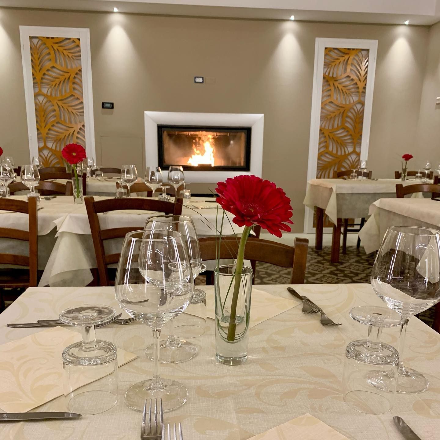 La sala da pranzo di un ristorante presenta tavoli con tovaglie bianche, bicchieri da vino e fiori rossi, disposti attorno a un accogliente camino.