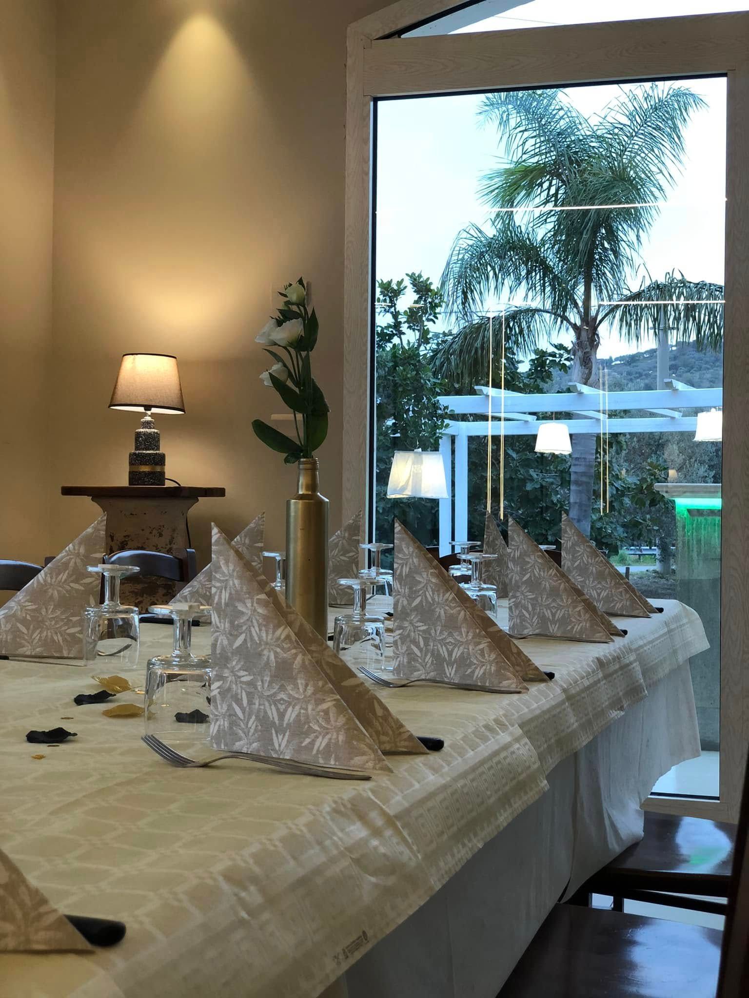 Una tavola da pranzo formale apparecchiata con tovaglioli piegati a fantasia e un vaso, affacciata su un patio con una palma al crepuscolo.