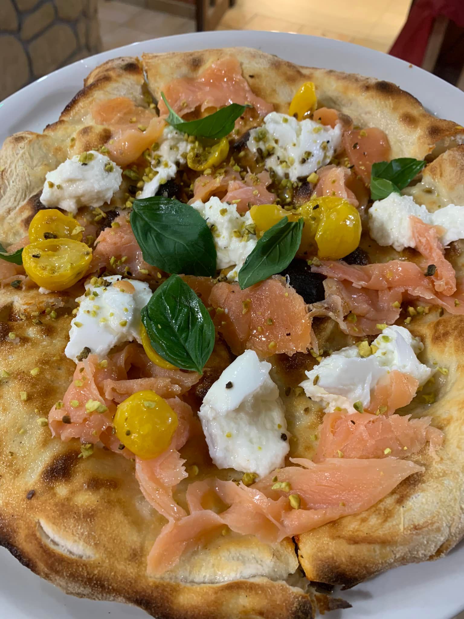 Una pizza gourmet condita con salmone affumicato, formaggio bianco, pomodorini gialli, basilico fresco e pistacchi tritati.