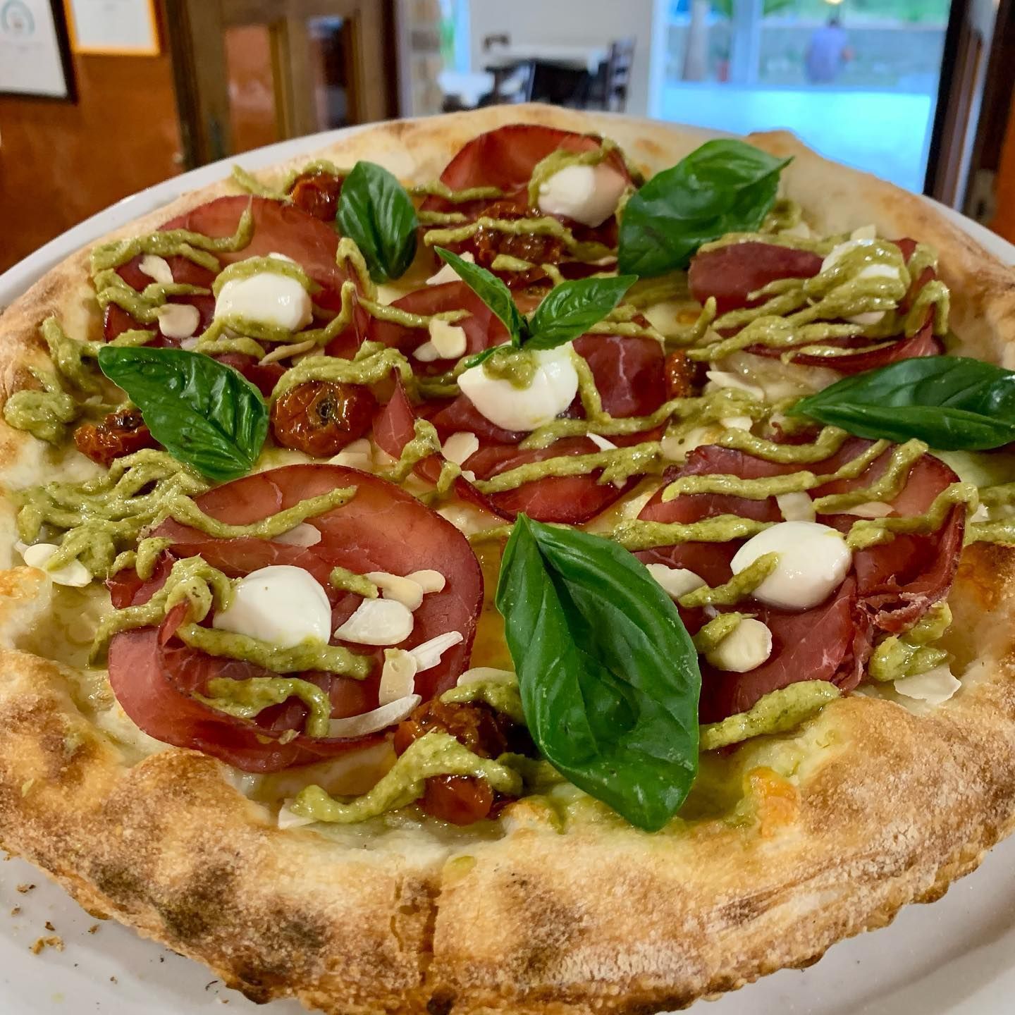 Una pizza condita con fette di salame, perle di mozzarella fresca, un filo di pesto, pomodori secchi e foglie di basilico.