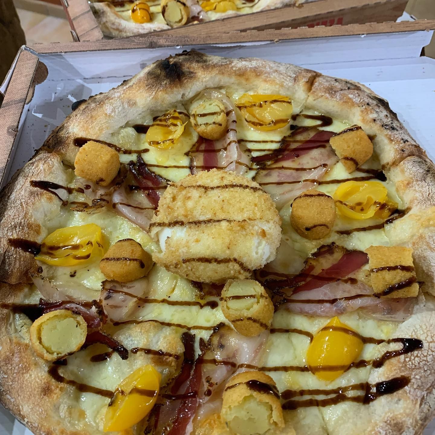 Una pizza guarnita con crocchette dorate, pomodorini gialli, pancetta, formaggio fuso e una spruzzata di glassa balsamica.