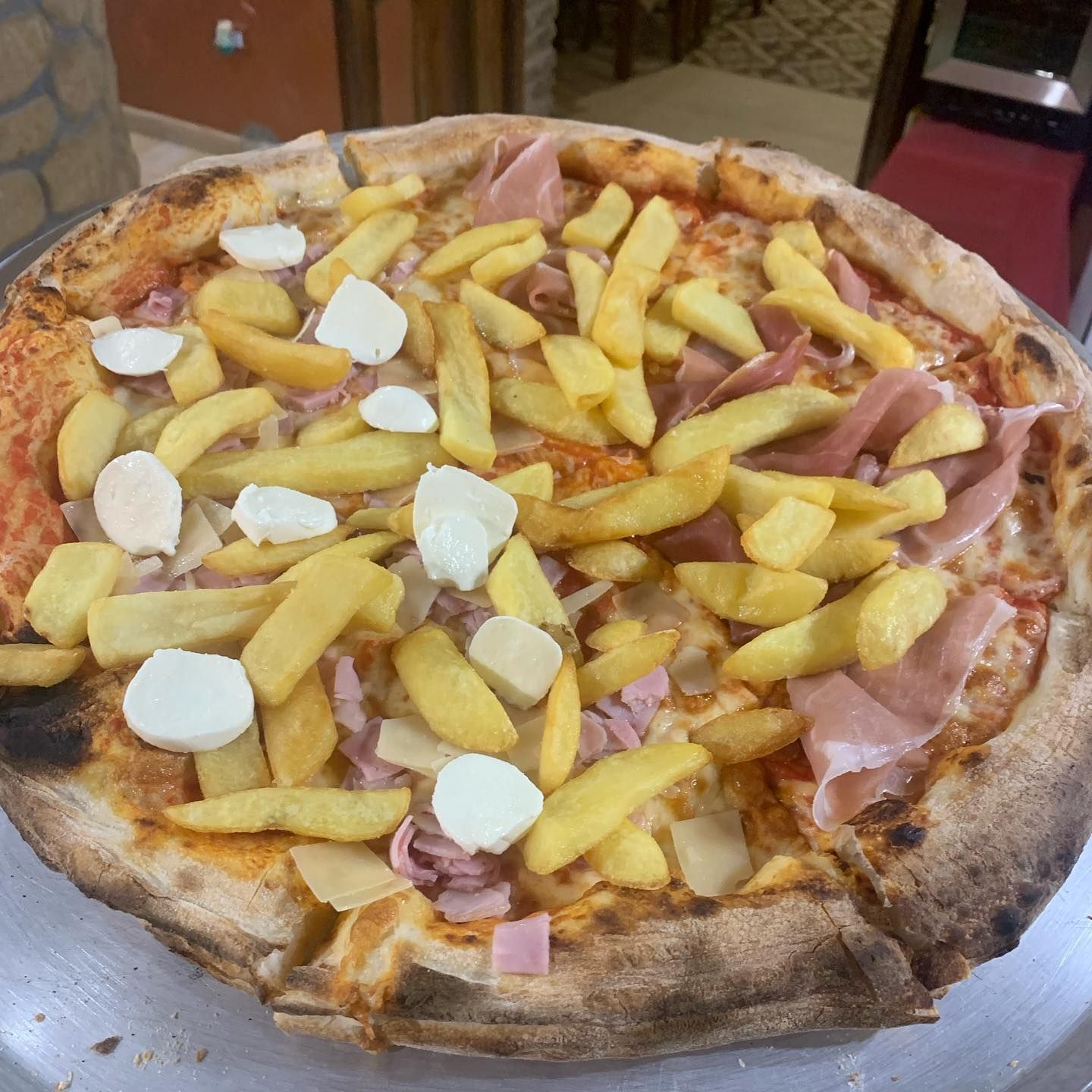 Una pizza condita con patatine fritte, fette di prosciutto e pezzi di mozzarella fresca, adagiata su un vassoio di metallo.