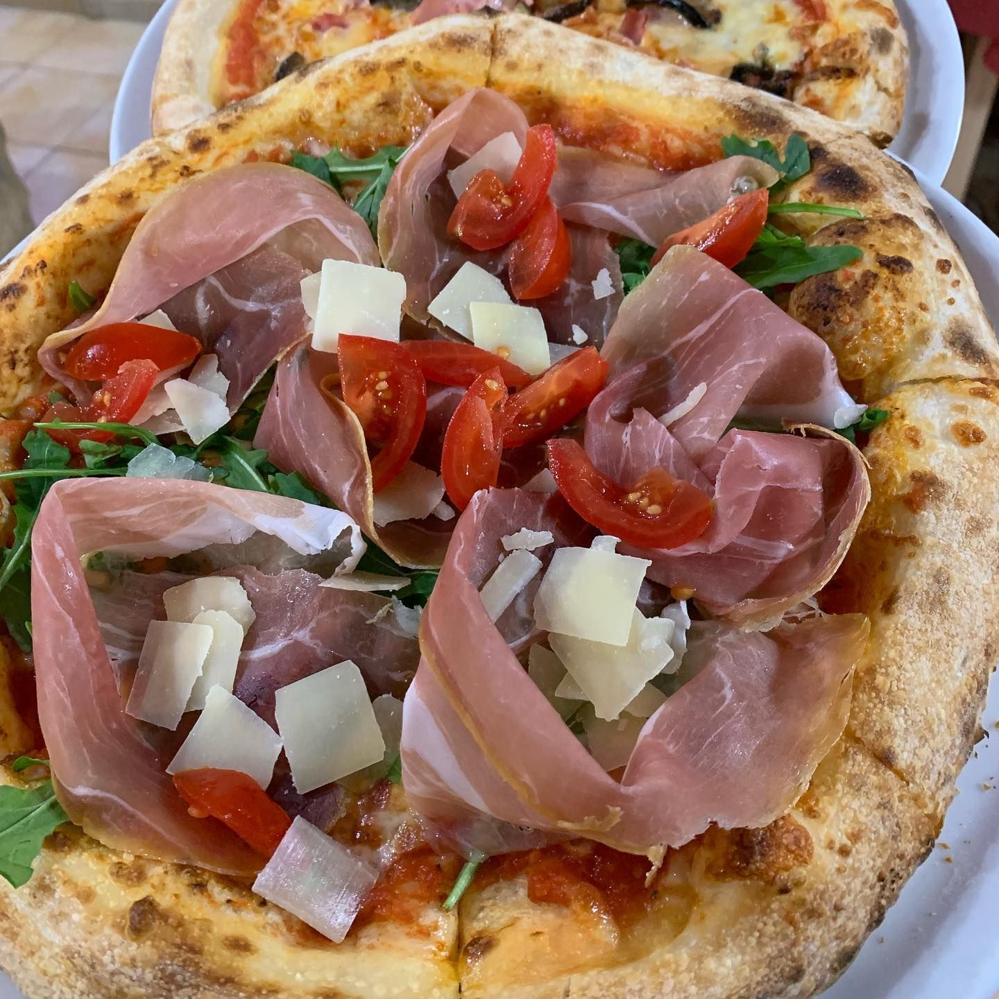 Una pizza condita con prosciutto crudo, rucola fresca, pomodorini e scaglie di parmigiano, servita su un piatto bianco.