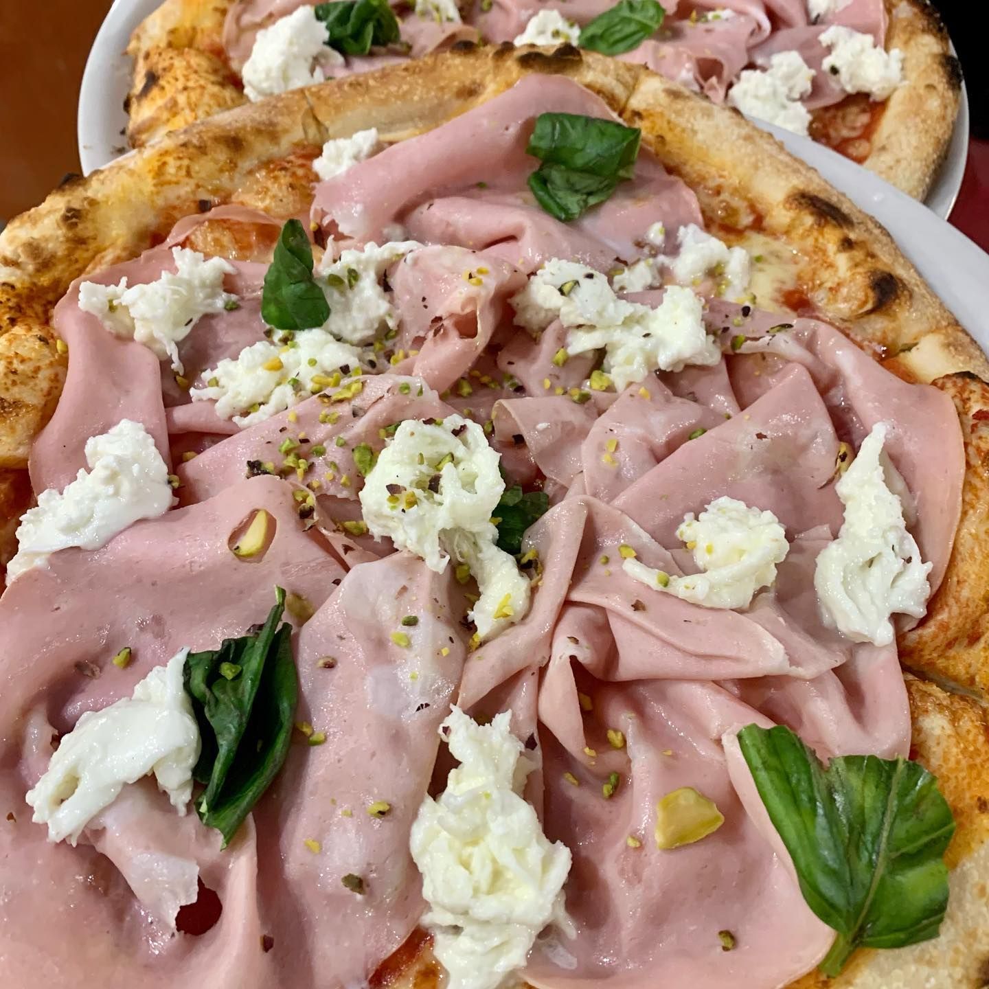 Due pizze condite con mortadella, mozzarella fresca, foglie di basilico e pistacchi tritati su una base sottile e dorata.