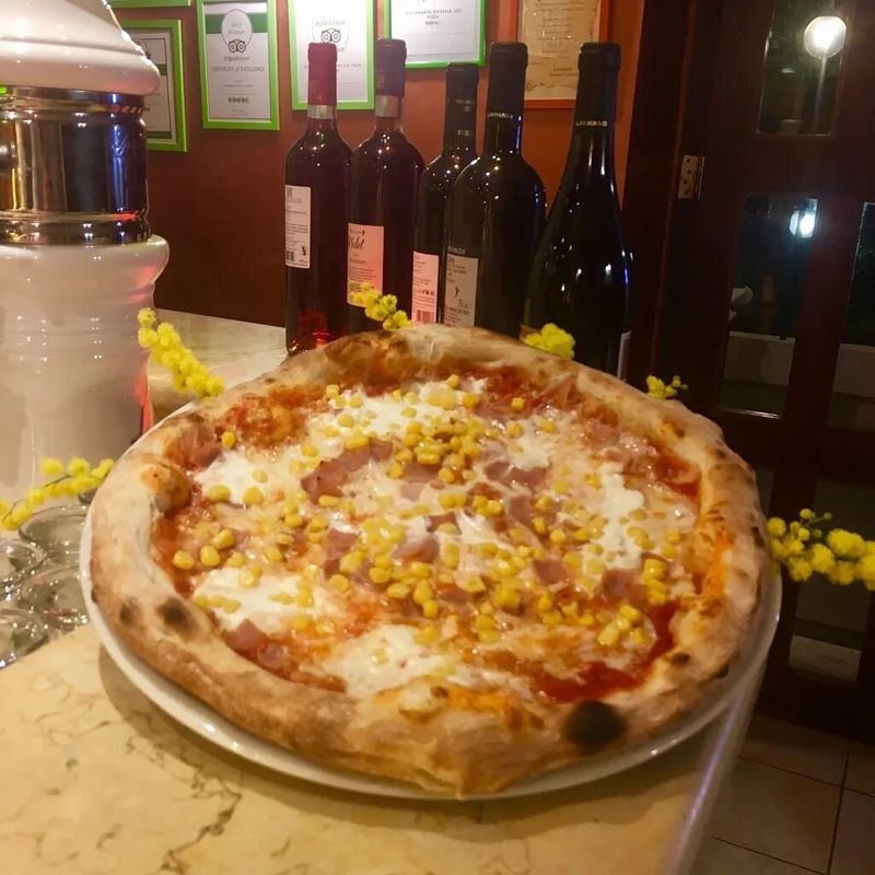 In un ristorante, su un tavolo davanti a diverse bottiglie di vino, si trova una pizza condita con prosciutto e mais.