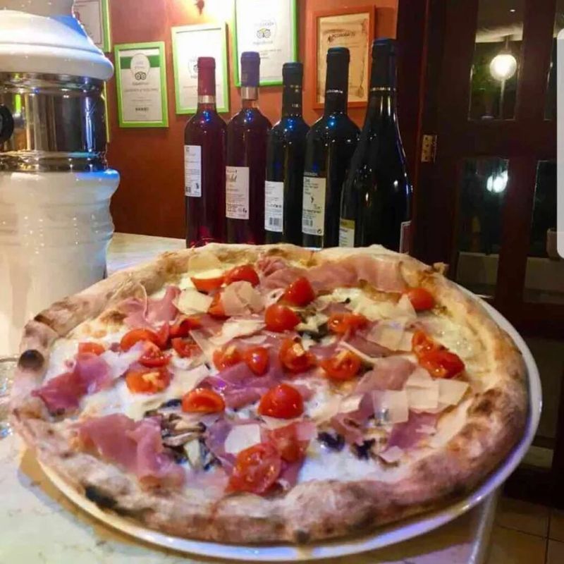 In un ristorante, su un tavolo davanti a diverse bottiglie di vino, si trova una pizza con pomodorini e prosciutto crudo.