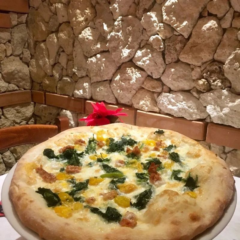 Una pizza gourmet condita con spinaci, pomodori gialli e formaggio, servita su un piatto bianco davanti a un rustico muro di pietra.