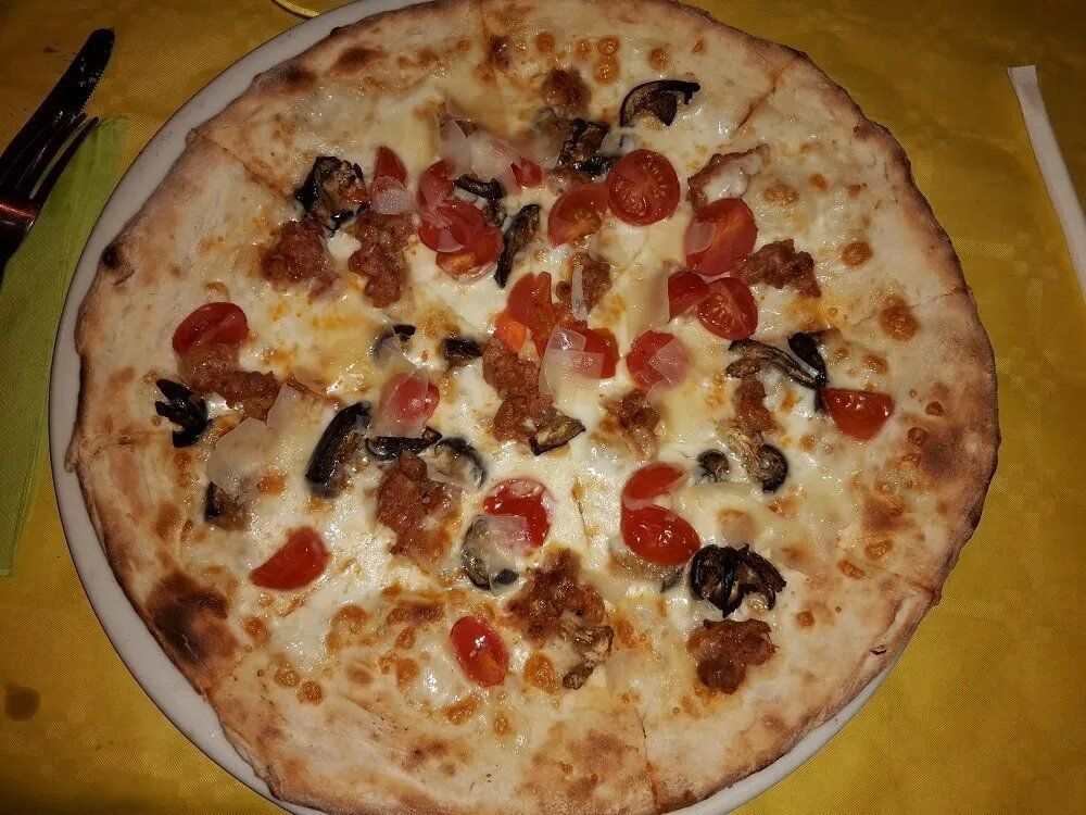 Una pizza a crosta sottile condita con pomodorini, salsiccia e funghi scuri, servita su un piatto bianco appoggiato su una tovaglia gialla.