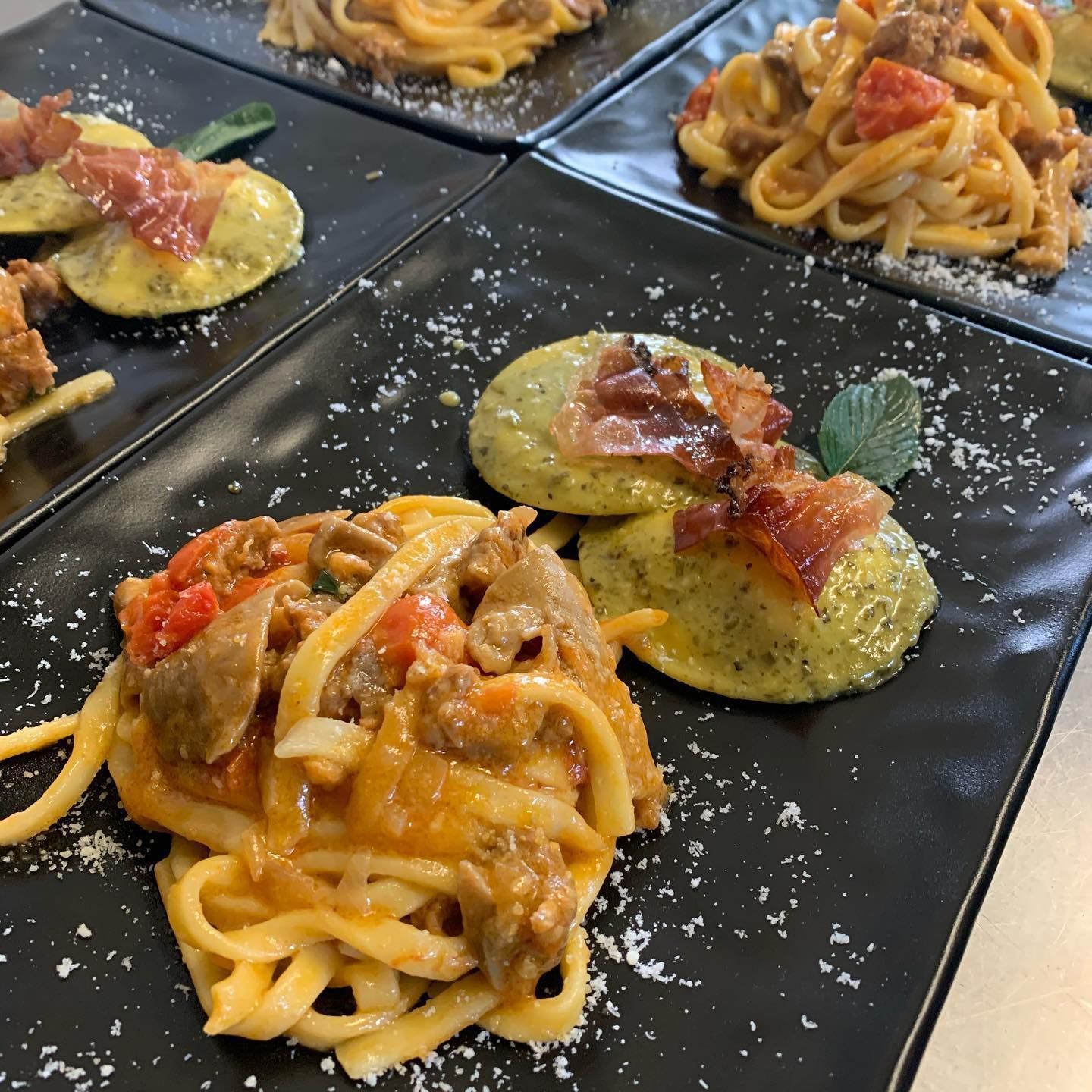 Quattro piatti rettangolari neri, ognuno con una porzione di pasta e due ravioli ripieni guarniti con prosciutto croccante.