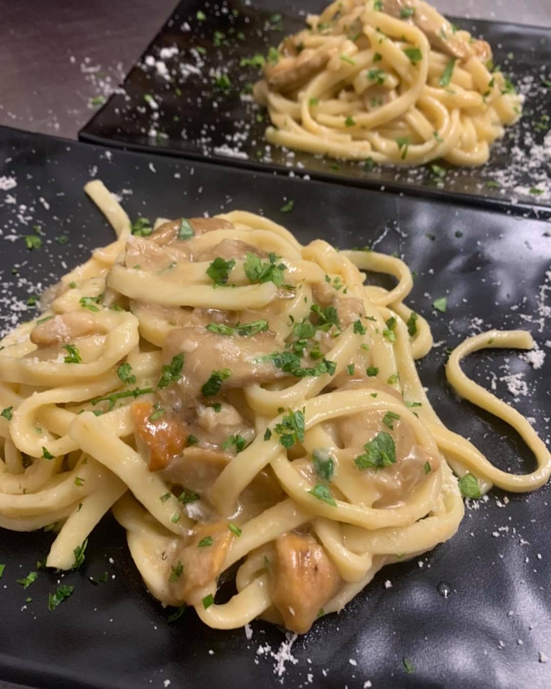 Due porzioni di pasta cremosa ai funghi, guarnita con prezzemolo tritato e formaggio grattugiato, servite su piatti rettangolari neri.