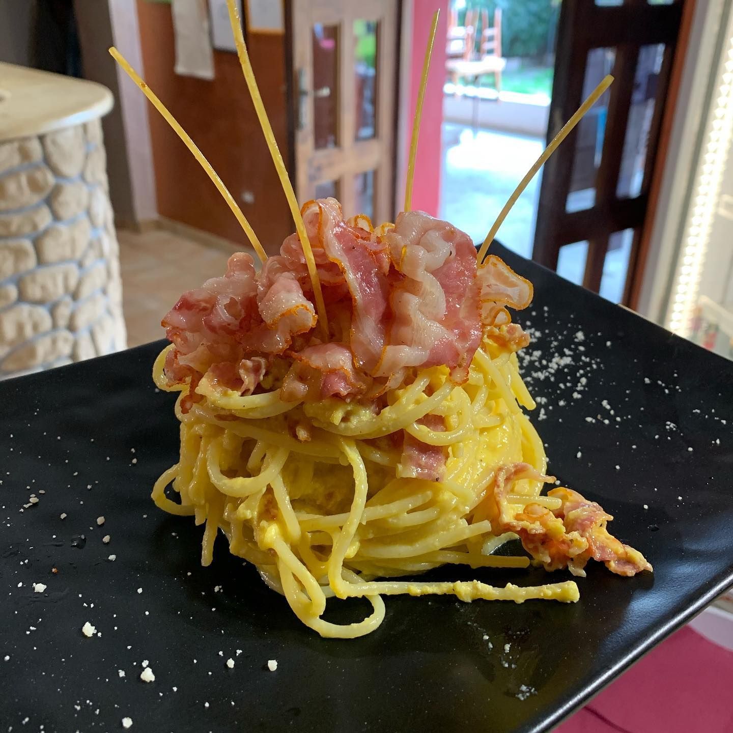 Una porzione di spaghetti alla carbonara con pancetta croccante su un piatto nero, decorato con fili di pasta cruda disposti verticalmente.