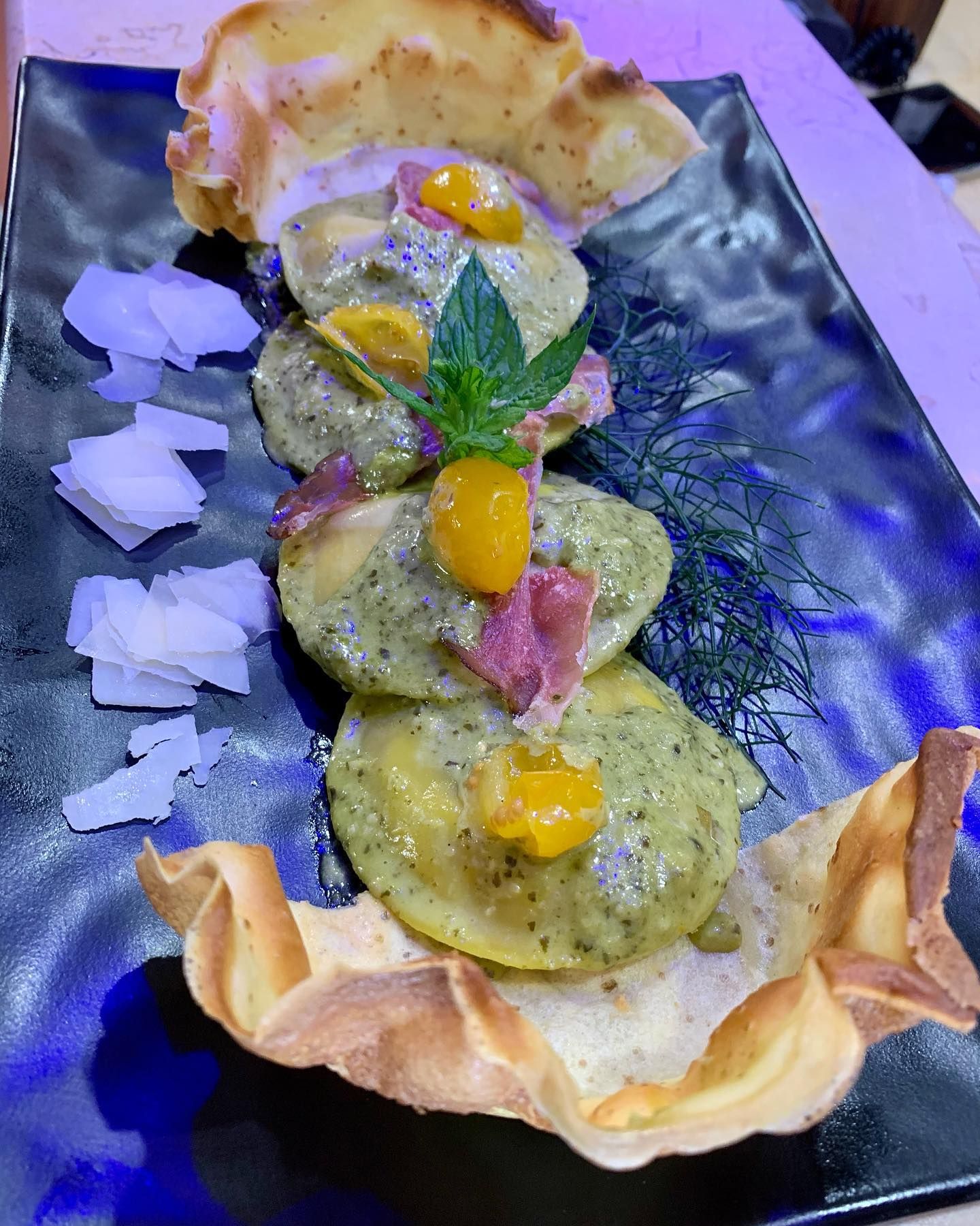 Una fila di ravioli al pesto guarniti con pomodorini, prosciutto e erbe aromatiche, serviti in una ciotola di pane croccante e dorata.