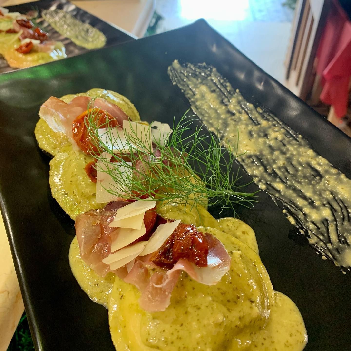 Un piatto di ravioli con una cremosa salsa verde, guarniti con sottili fette di prosciutto, pomodori secchi e finocchio.