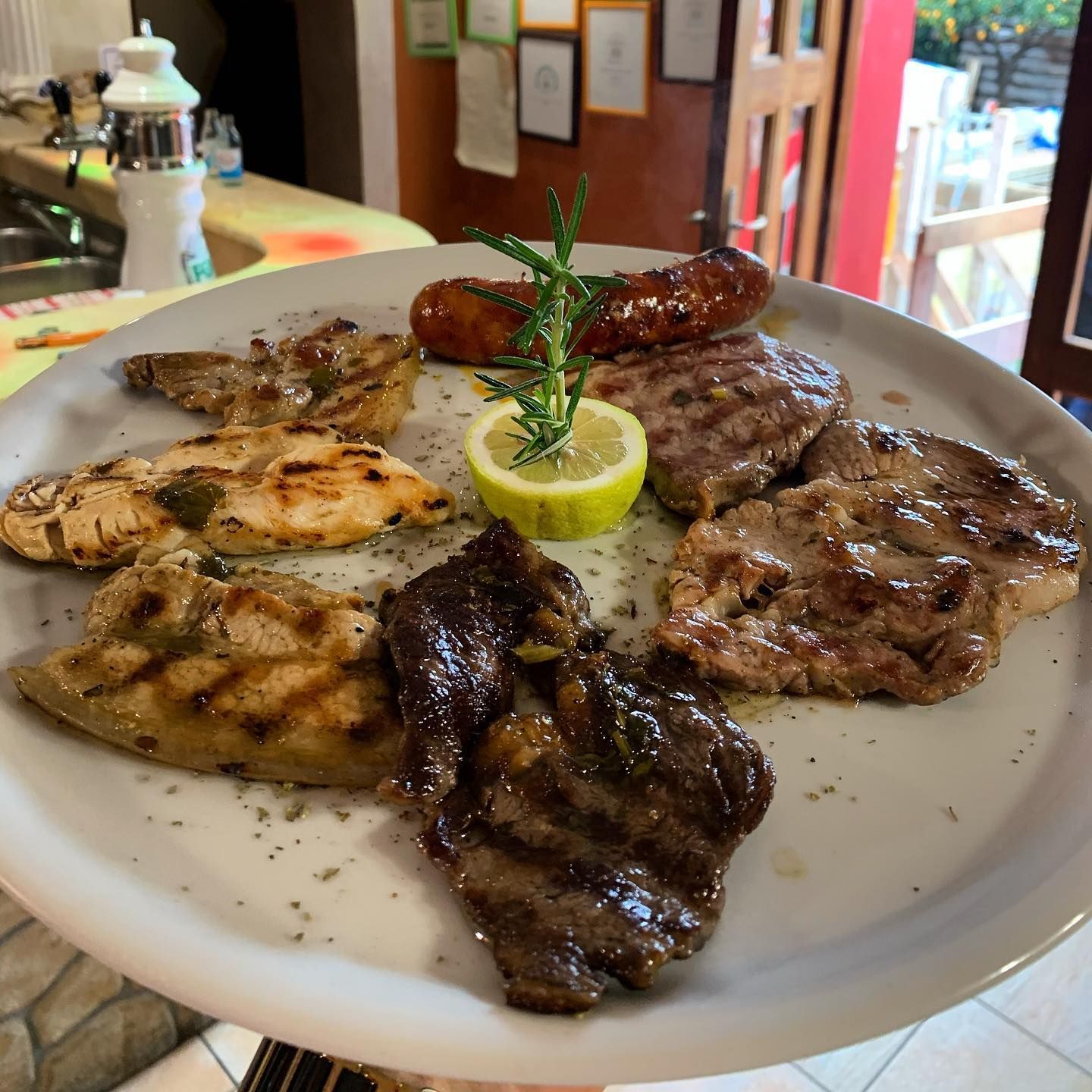 Un piatto bianco su cui sono disposti diversi tipi di carne grigliata, una salsiccia e uno spicchio di limone guarnito con un rametto di rosmarino.