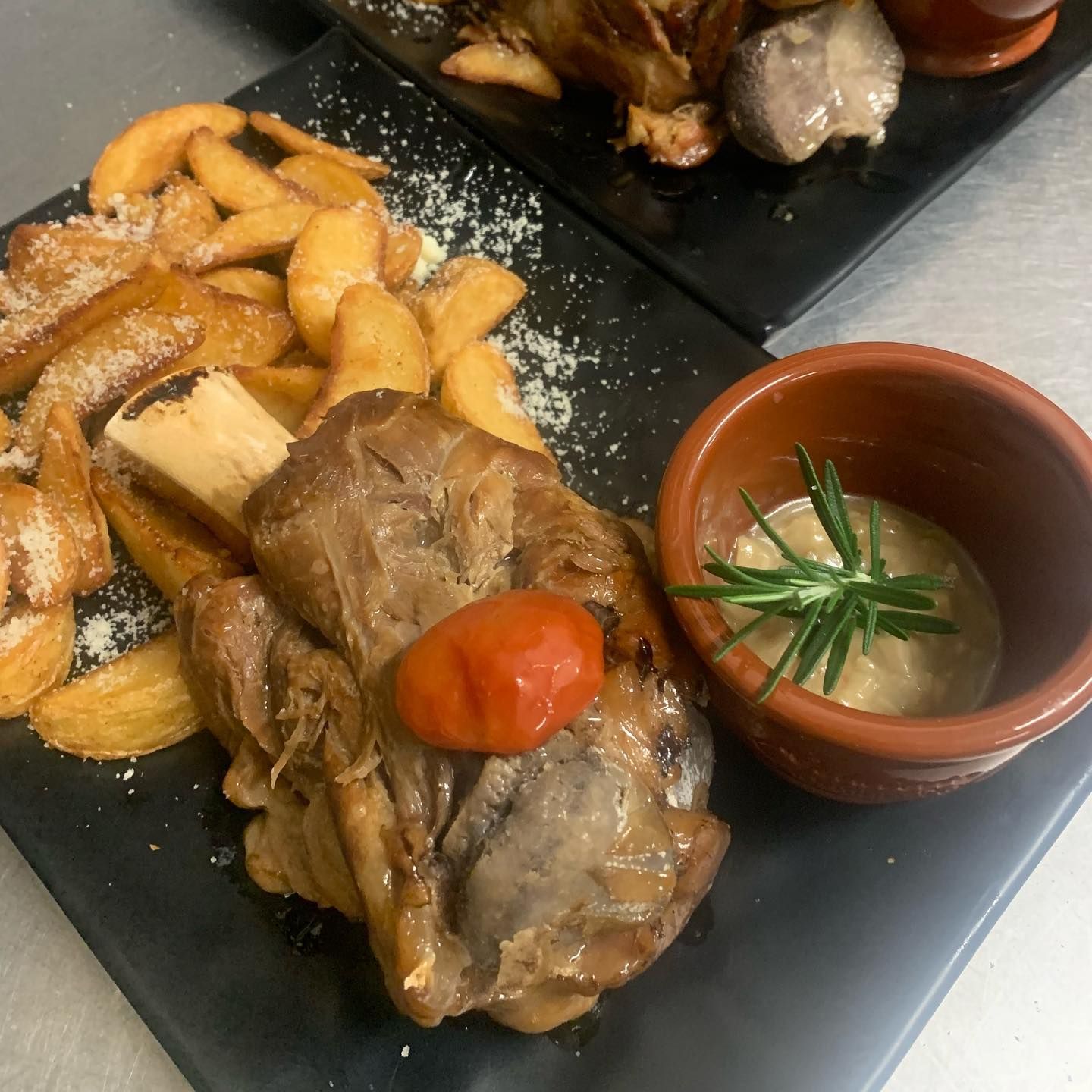 Un piatto con uno stinco di maiale arrosto, spicchi di patate cosparsi di formaggio, un pomodorino ciliegino e una ciotolina di salsa.
