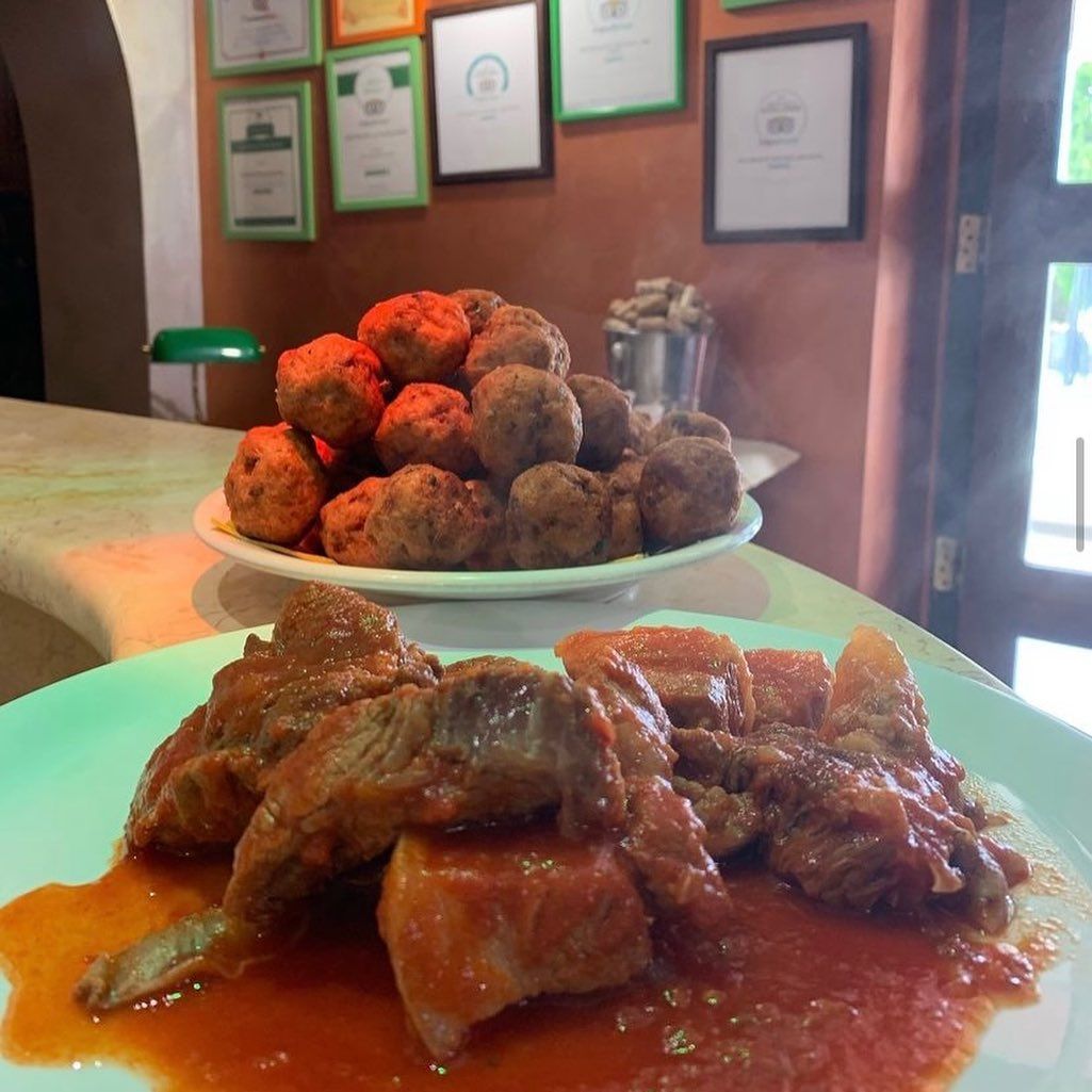 In primo piano un piatto di carne al sugo di pomodoro, con un mucchio di polpette su un piatto alle sue spalle.