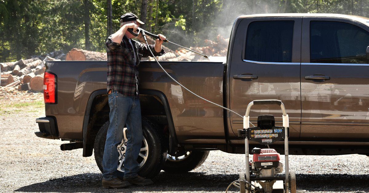 A man using a power washer to clean the inside of his truck bed. The man is wearing a hat.