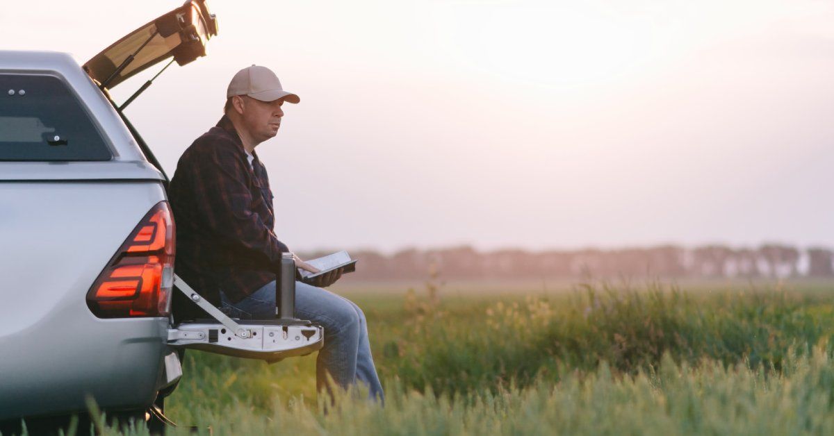 A man sitting on the end of his truck bed in a grassy field. He looks into the distance, a book in his hands.