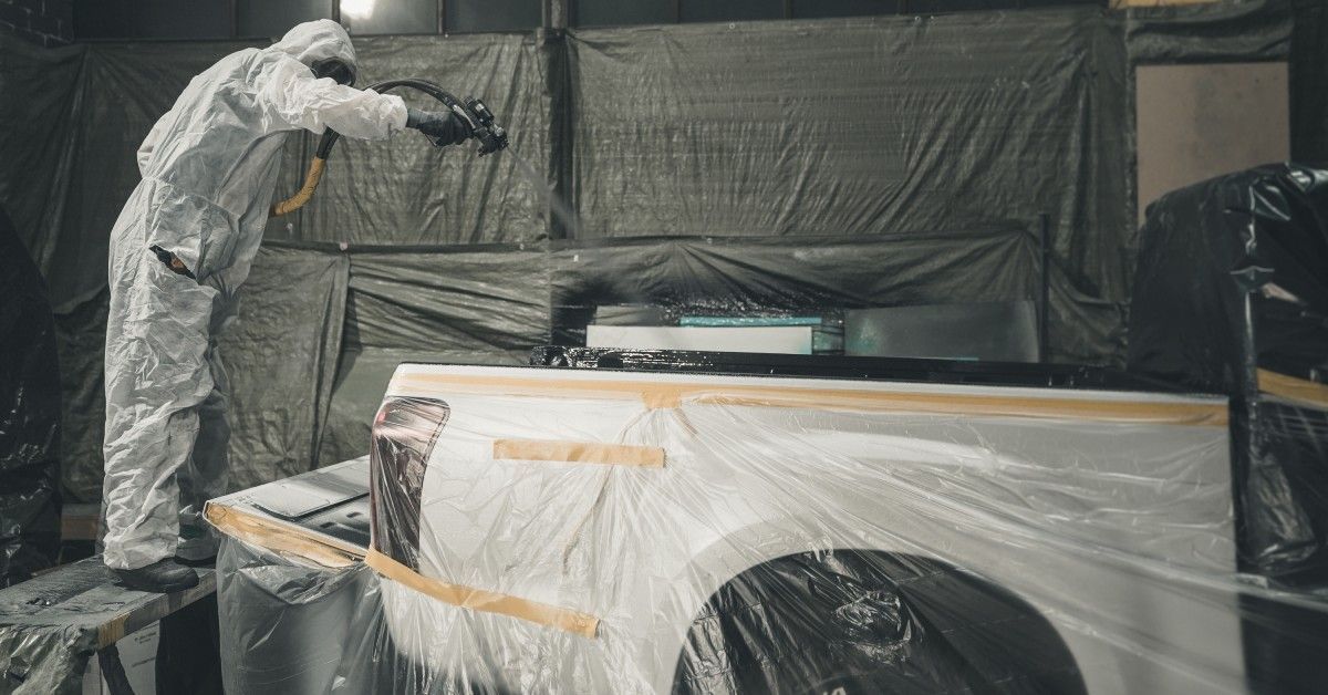 A professional mechanic in protective clothing applies a truck bed liner in a controlled environment indoors.