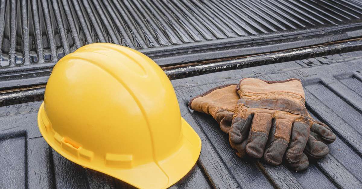 A clean, black pickup truck bed is open. A yellow hard hat and two brown gloves sit in an otherwise empty truck bed.