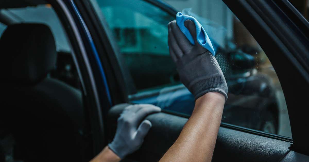 A person wearing gloves is using a small blue rag to wipe the interior of a tinted car window clean.