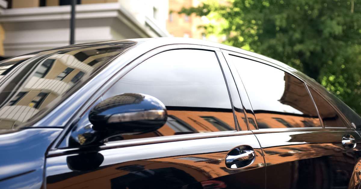 A sleek black car with dark, tinted windows is parked outside on a sunny day near a large green tree