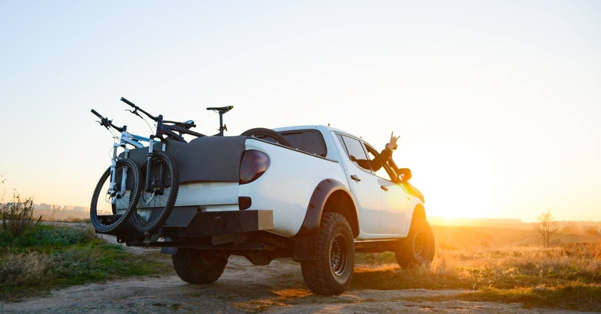 A white pickup truck is parked on an off-road trail. The truck bed has two bikes that are hanging over the backdoor.