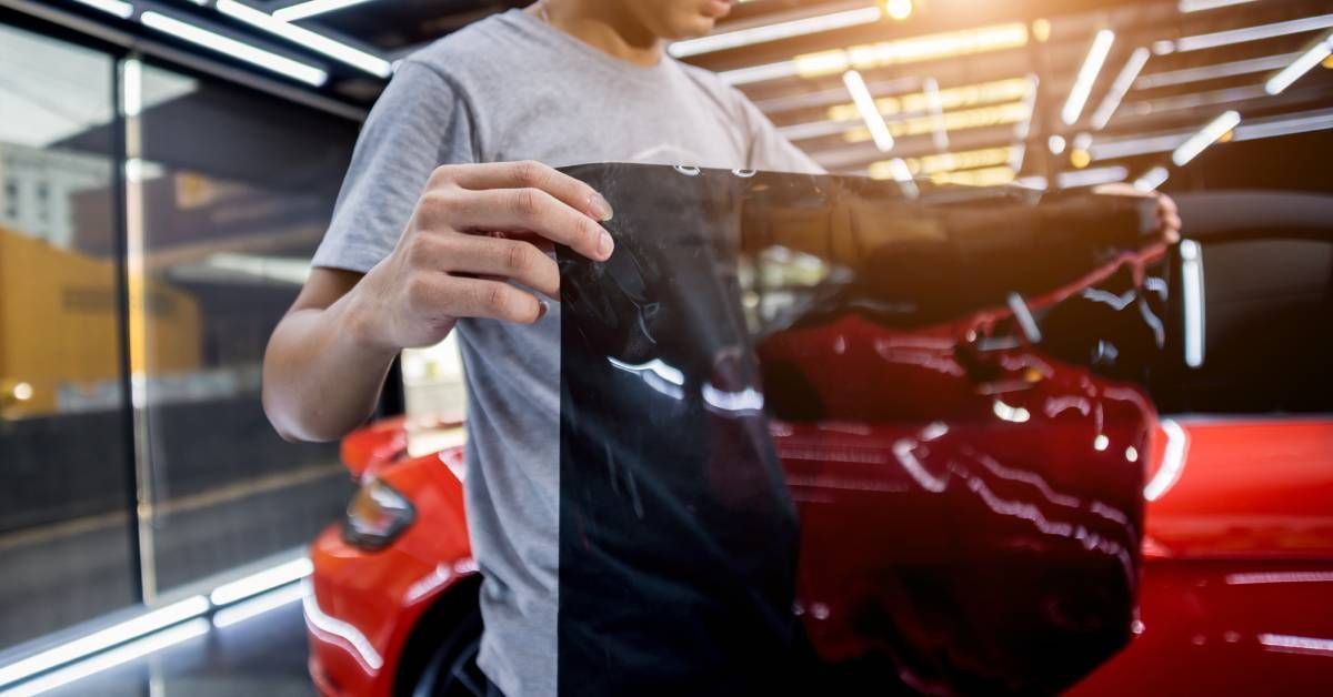 A person wearing a gray short-sleeved shirt is holding a black foil for window tinting while standing next to a red car.