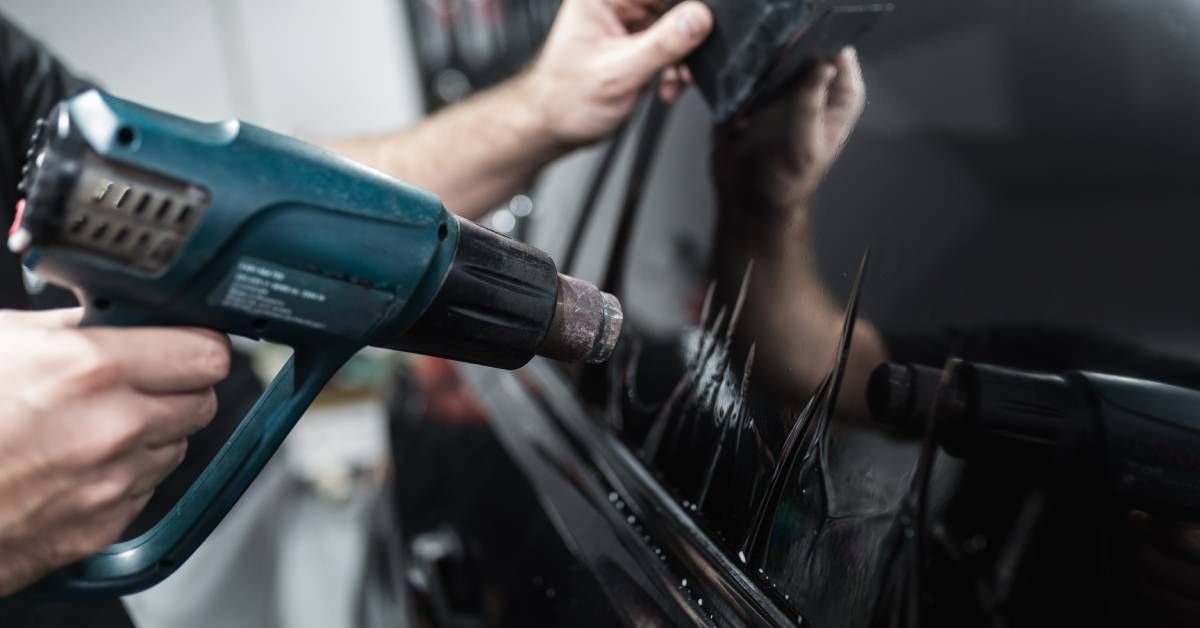 An employee is holding a blue tool and using it to apply a window tint to a black car inside a garag