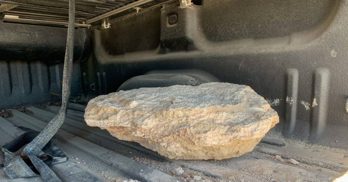 A pickup truck carrying a large boulder in its truck bed. The bed is covered in dust and a strap is hanging from above.