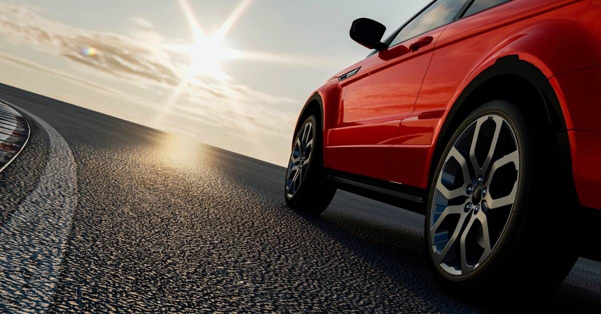 A shiny red car is driving on the road while the sun shines down from above, reflecting off the car’