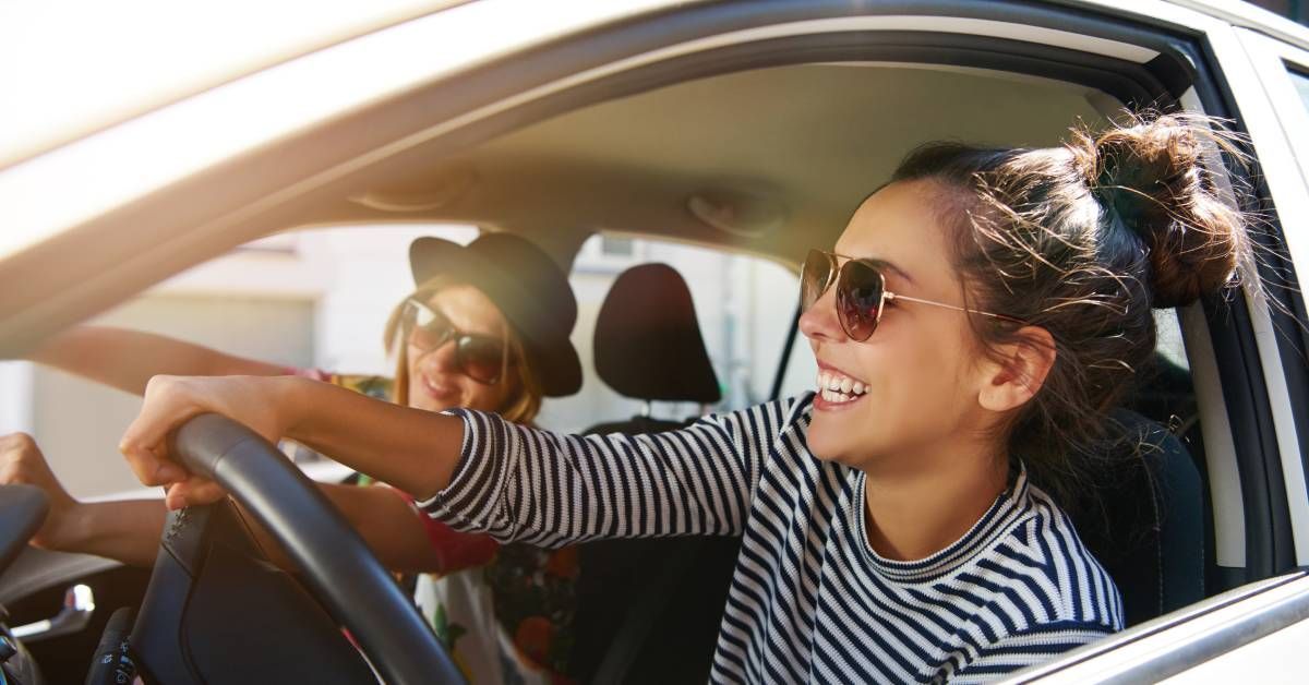 Two people are smiling together while wearing stylish dark sunglasses and driving in a car on a sunny day.