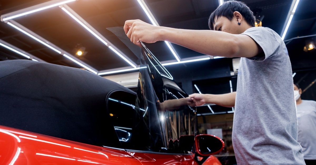 Two people in an automotive shop standing near a red car. One of the shop employees applies a tint to the car’s window.