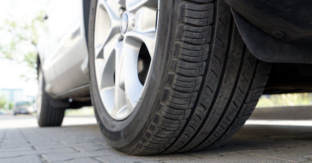 A car tire attached to a gray vehicle. The vehicle is parked in the shade and a tree is visible in the background.