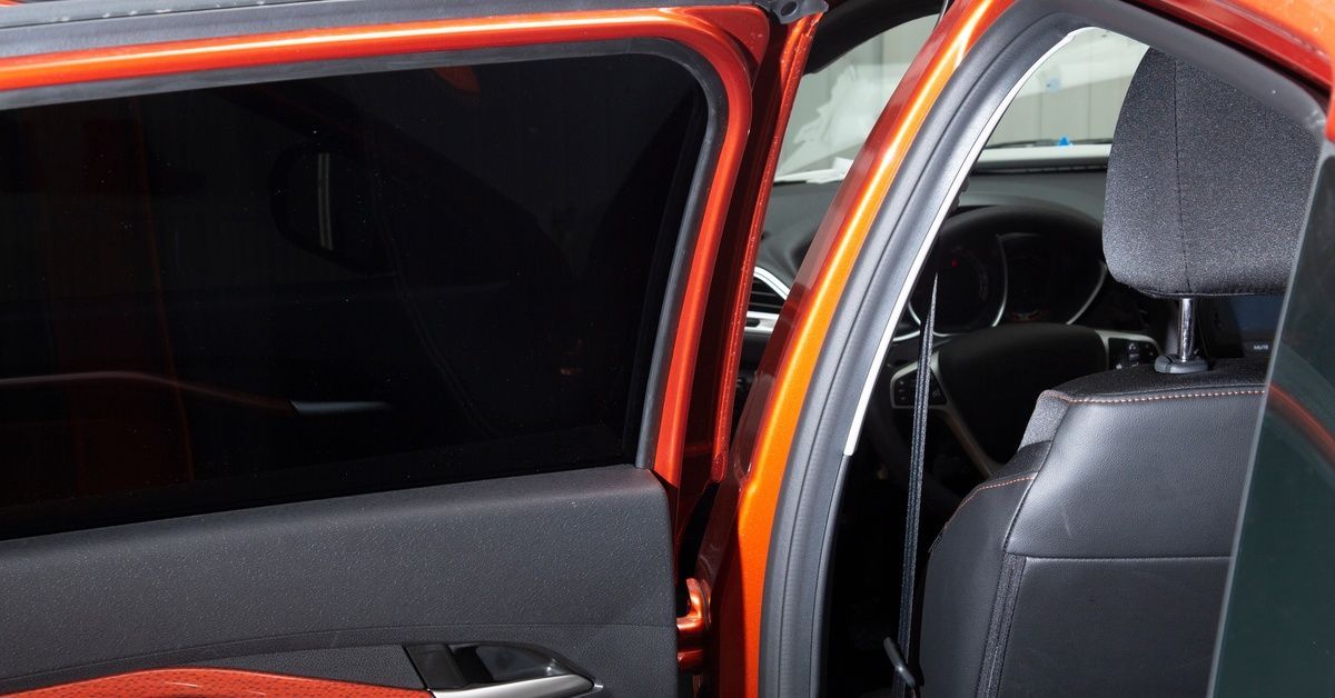 An orange car with dark tinted windows is parked near a white wall. One of the car’s back doors is wide open.