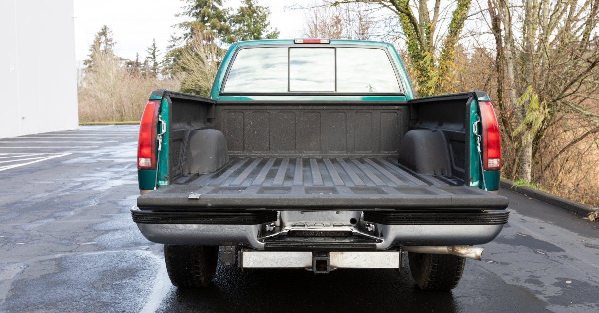 A blue pickup truck parked in an empty parking lot with its truck bed open. The parking lot is surrounded by trees.