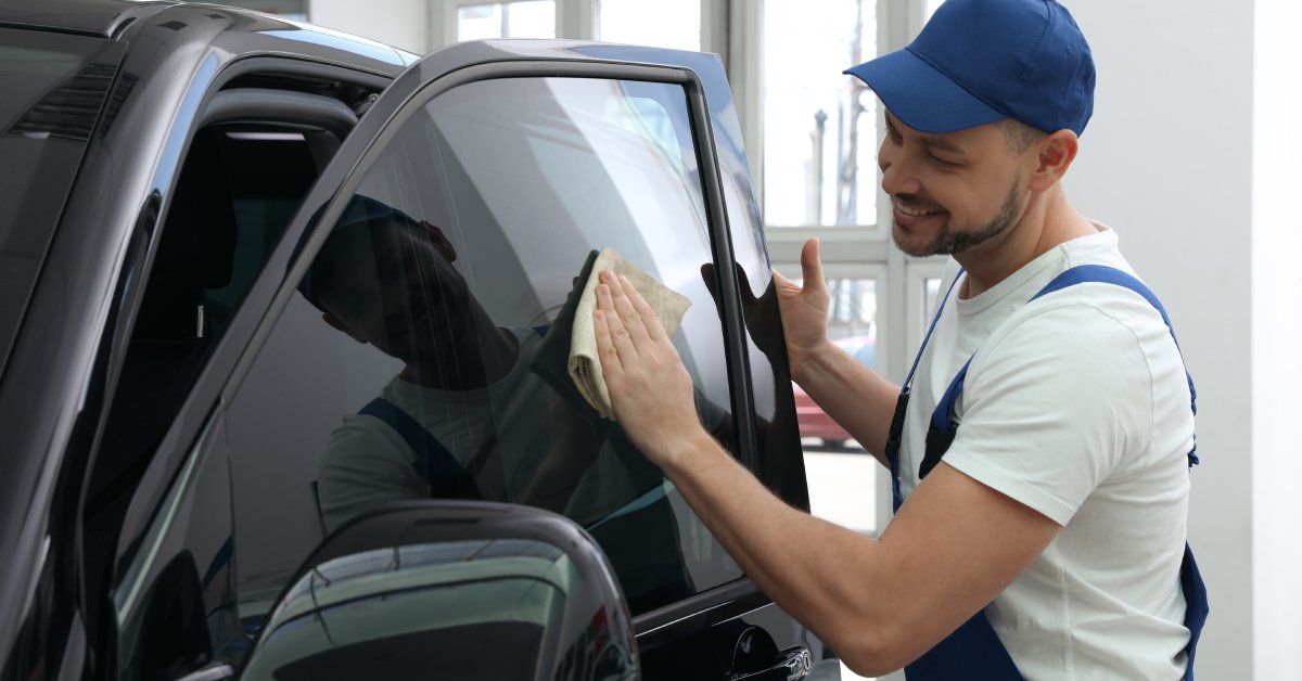 A person wearing a blue hat and blue overalls smiling while wiping a car window. The car window is tinted.