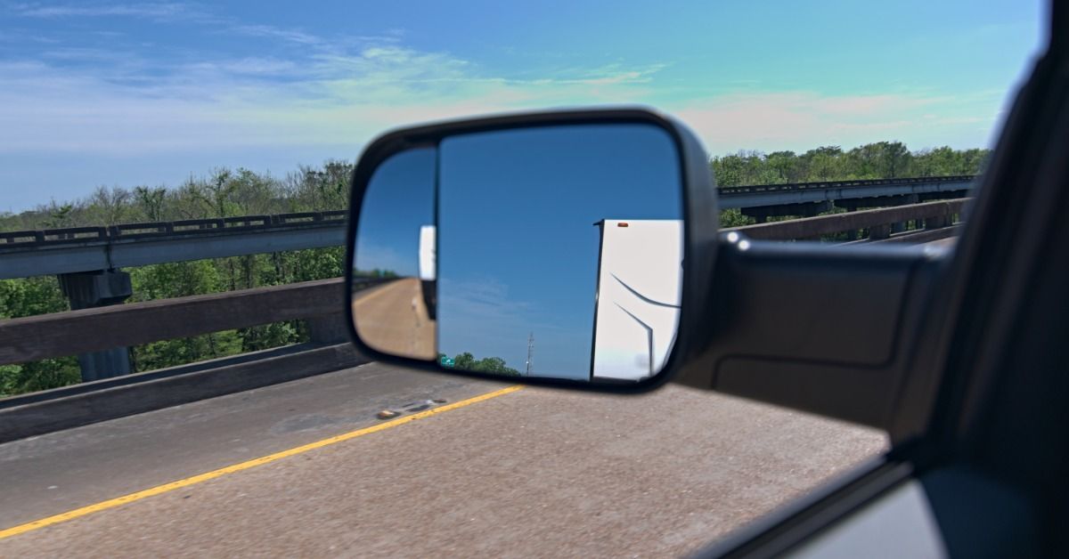 A towing mirror attached to the driver’s side of a truck towing a white trailer on a highway during the daytime.