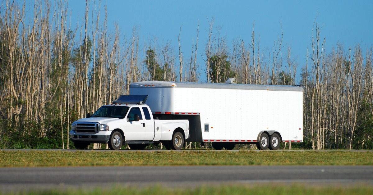 A white truck towing a trailer next to trees with very few leaves. The truck is parked on the side of the road.