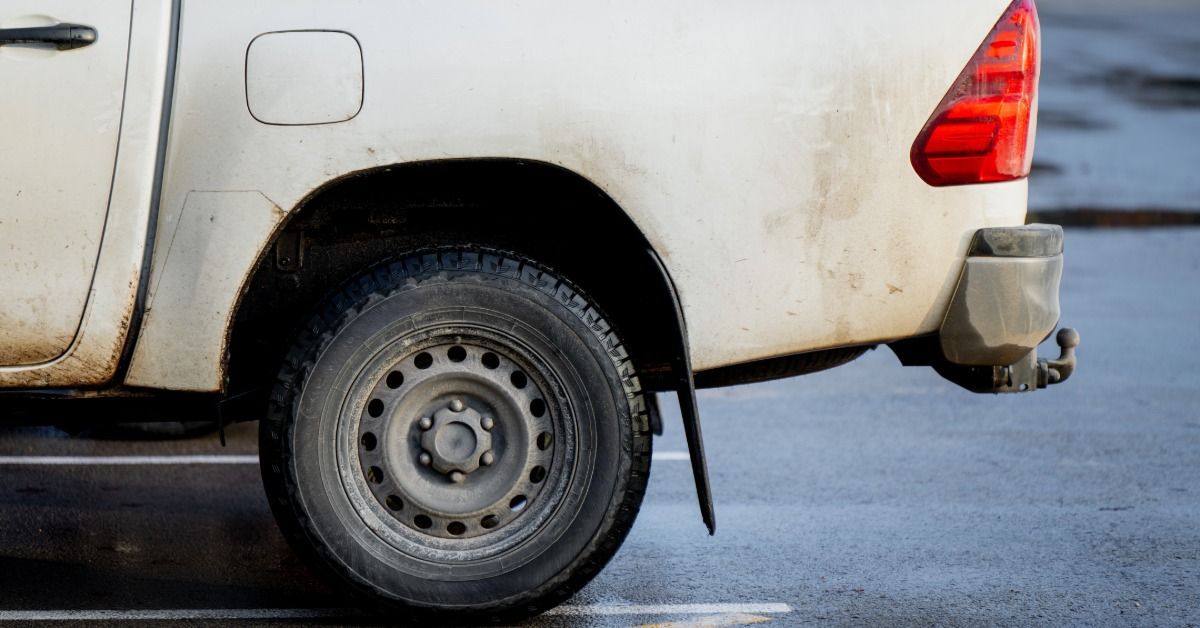 A white truck parked outside during the daytime. The truck bed door is up and dirt smudges are near the left taillight.