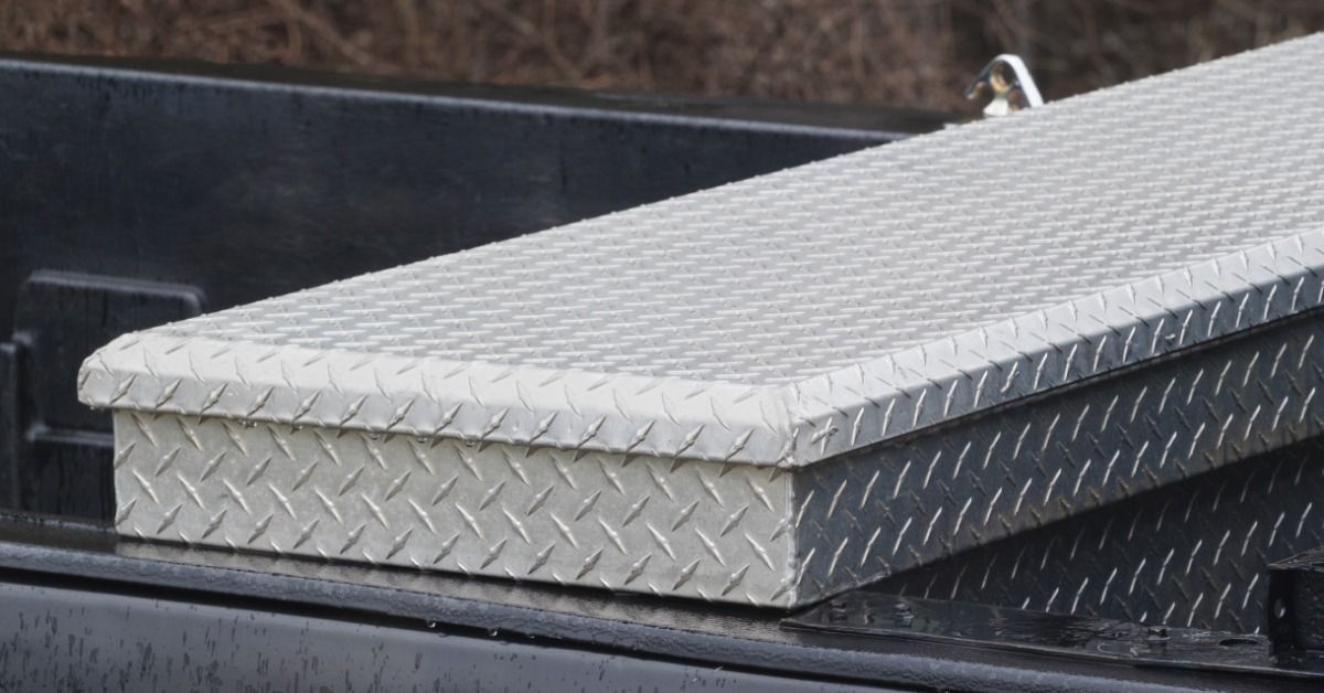 A gray vehicle parked outside near bushes during the daytime. A long metal toolbox is sitting in the truck bed.