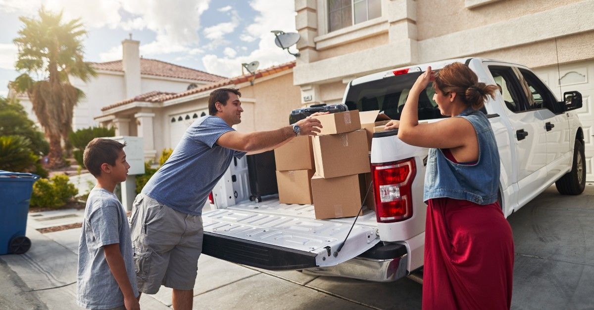 One parent packs boxes into their pickup truck bed while their child and significant other stand nearby and watch.