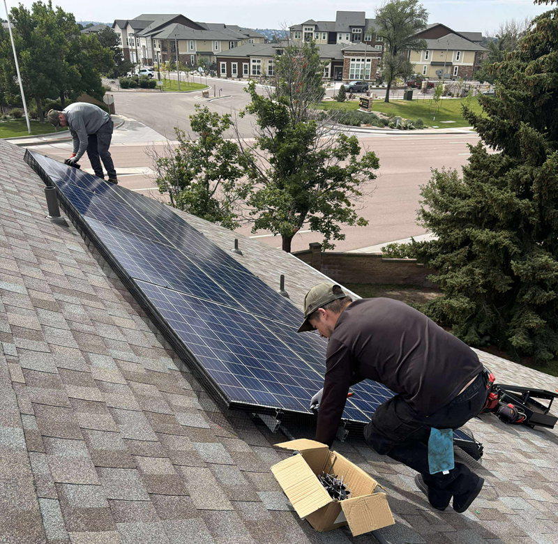 Two workers install solar panels on a residential roof, with a box of parts and houses in the background.