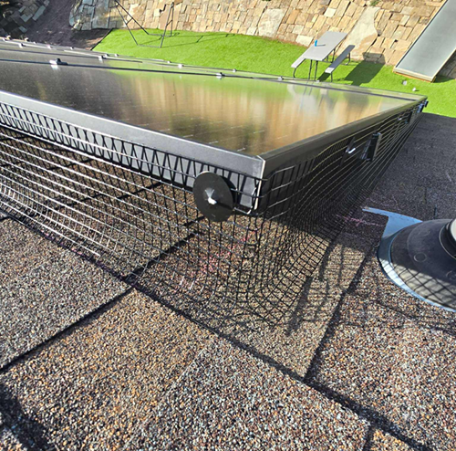 Solar panels on a shingled roof, secured with wire mesh. Overlooking a small green lawn and rock wall.